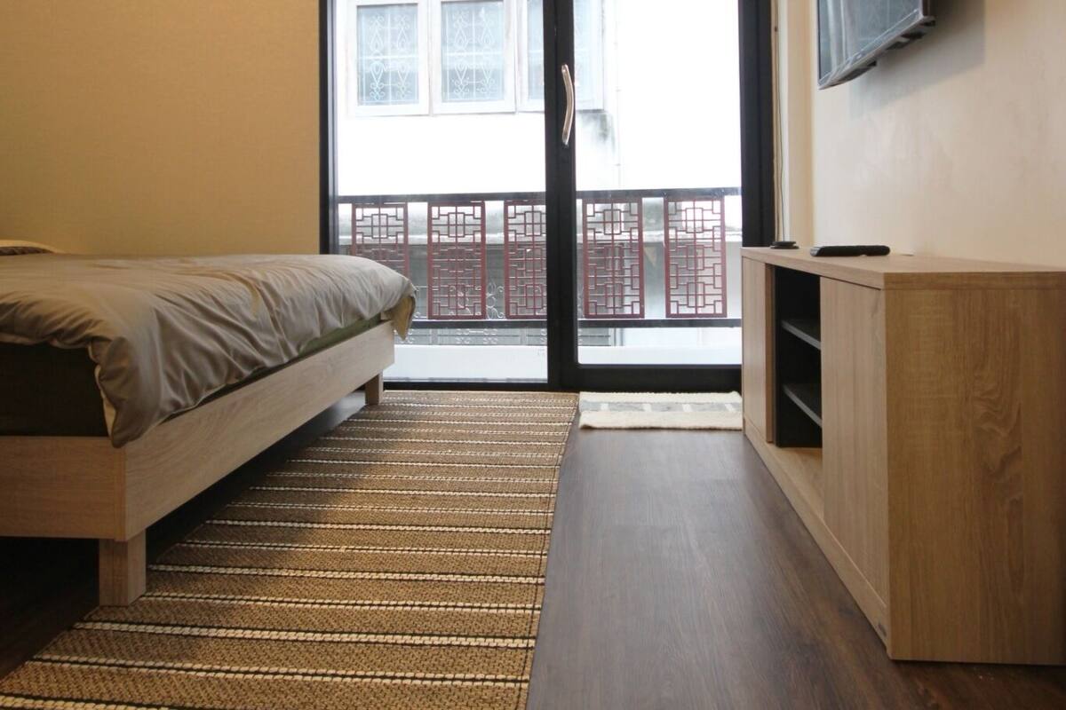 A bedroom with a bed, a TV mounted on the wall, and a sliding glass door leading to a balcony.