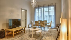 A living room with a TV, wooden furniture, a chandelier, and a large window with curtains. - Baratero City II Apartment (Sofia)