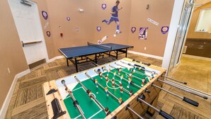 A room with a foosball table, a ping pong table, and a wall with sports-themed decorations.