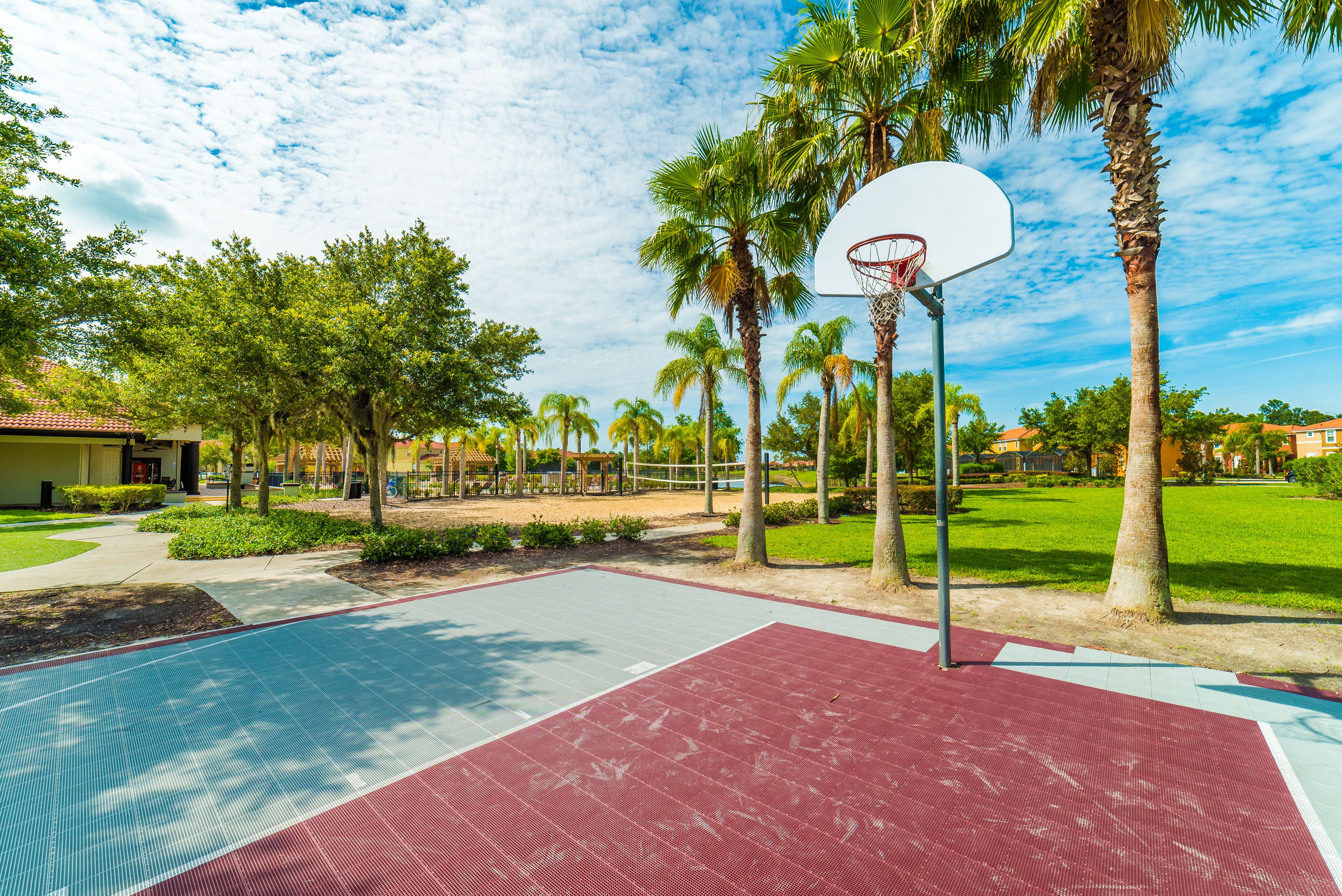 An outdoor basketball court with a red and blue playing surface, surrounded by palm trees and a clear sky.
