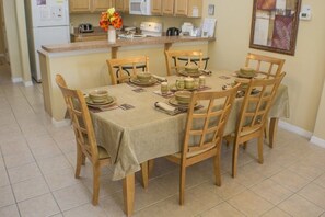 A dining area with a table set for four, surrounded by wooden chairs. A kitchen with a refrigerator, microwave, and toaster is visible in the background.