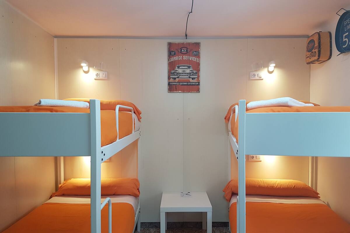 Quarto tipo dormitório com duas beliches, uma mesinha e um cartaz na parede.