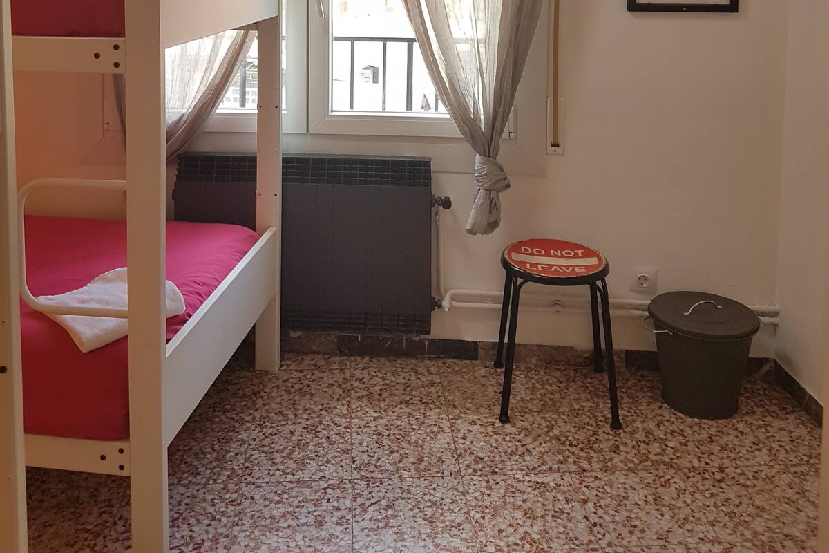 Um quarto de dormitório com beliches, um radiador, uma janela com cortinas, um cartaz emoldurado e um banquinho pequeno.