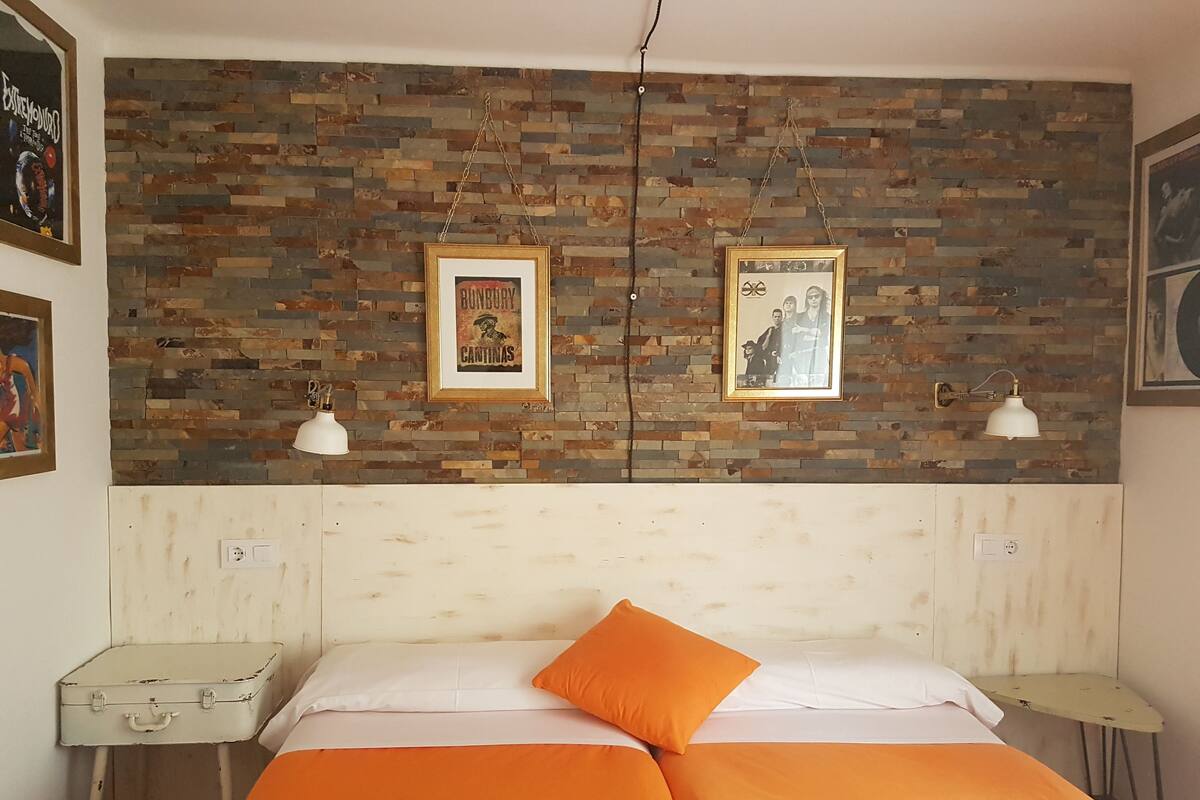 Quarto com parede em padrão tijolo, duas imagens emolduradas, cama com roupa de cama laranja e uma pequena mesa com luminária.