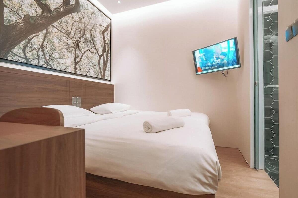 A modern hotel room with a wooden headboard, a large bed with white linens, a flat-screen TV mounted on the wall, and a glass door leading to a shower area.