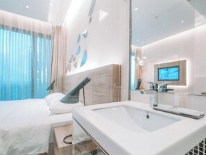 A modern hotel room with a large bed, a glass shower enclosure, a white sink, and a flat-screen TV mounted on the wall. - L Hotel @ Bugis Ville (Singapore)