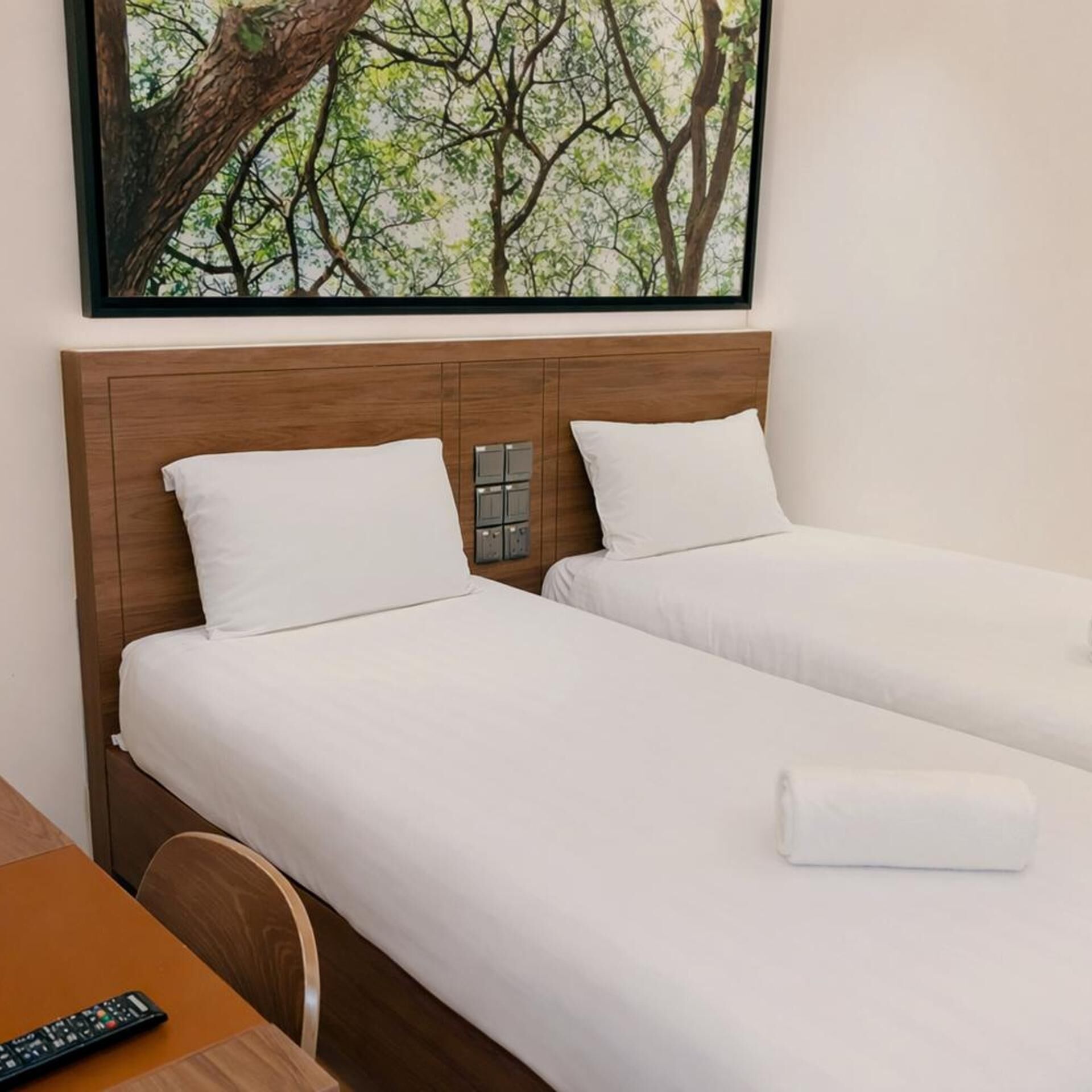Une chambre d’hôtel avec deux lits simples, une tête de lit en bois, un tableau représentant des arbres et une télécommande sur la table.