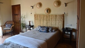 A bedroom with a large bed, a wooden door, a wicker chair, a small table, and a wall-mounted light fixture. - The African House (Tolox)