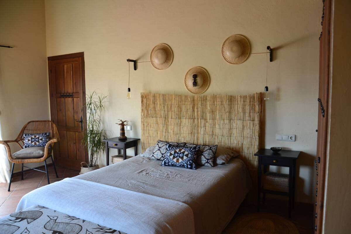Un dormitorio con una cama grande, una puerta de madera, una silla de mimbre, una mesita y una lámpara fijada a la pared.