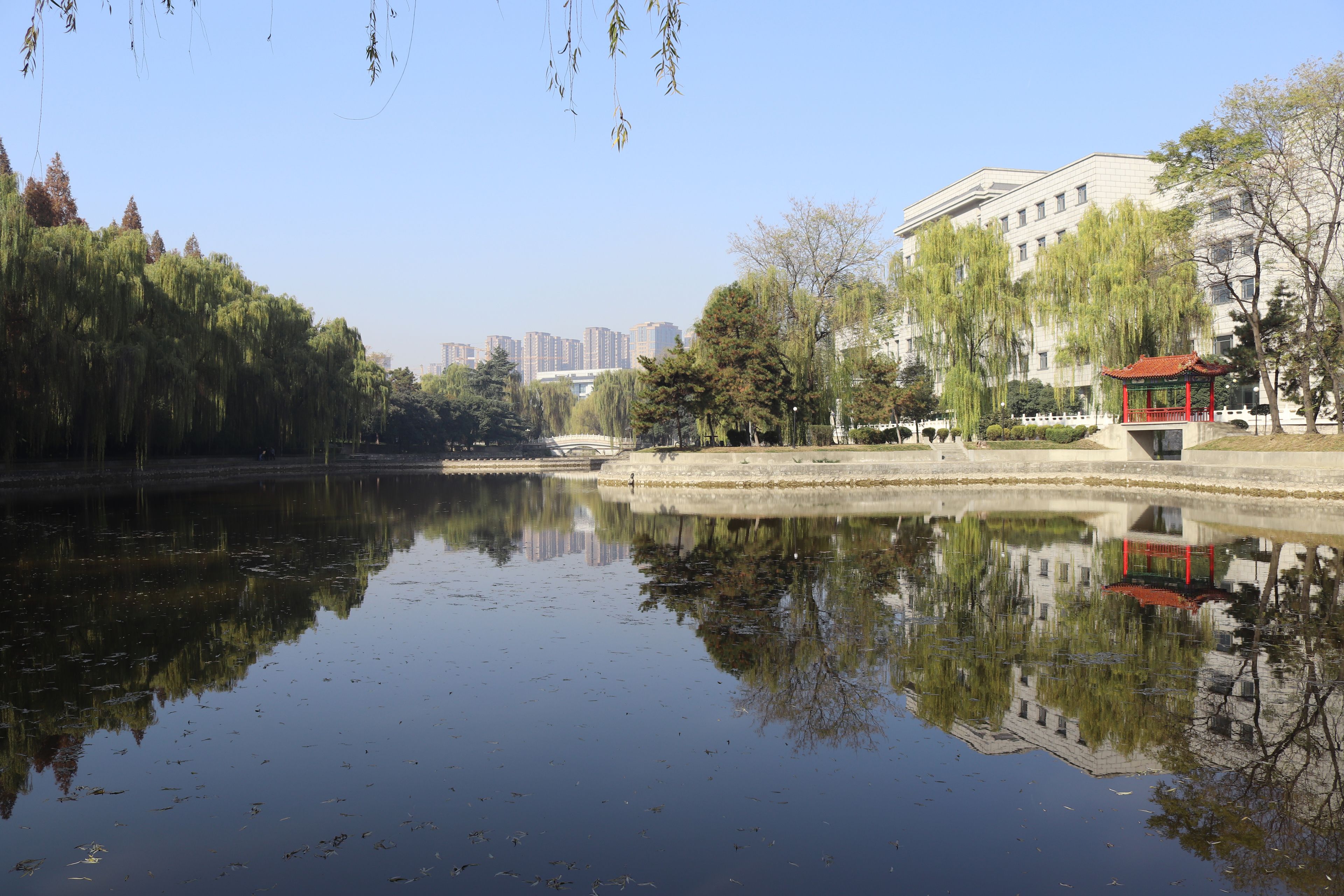 A calm lake with reflections of trees and buildings, a red pavilion, and a bridge in the background.