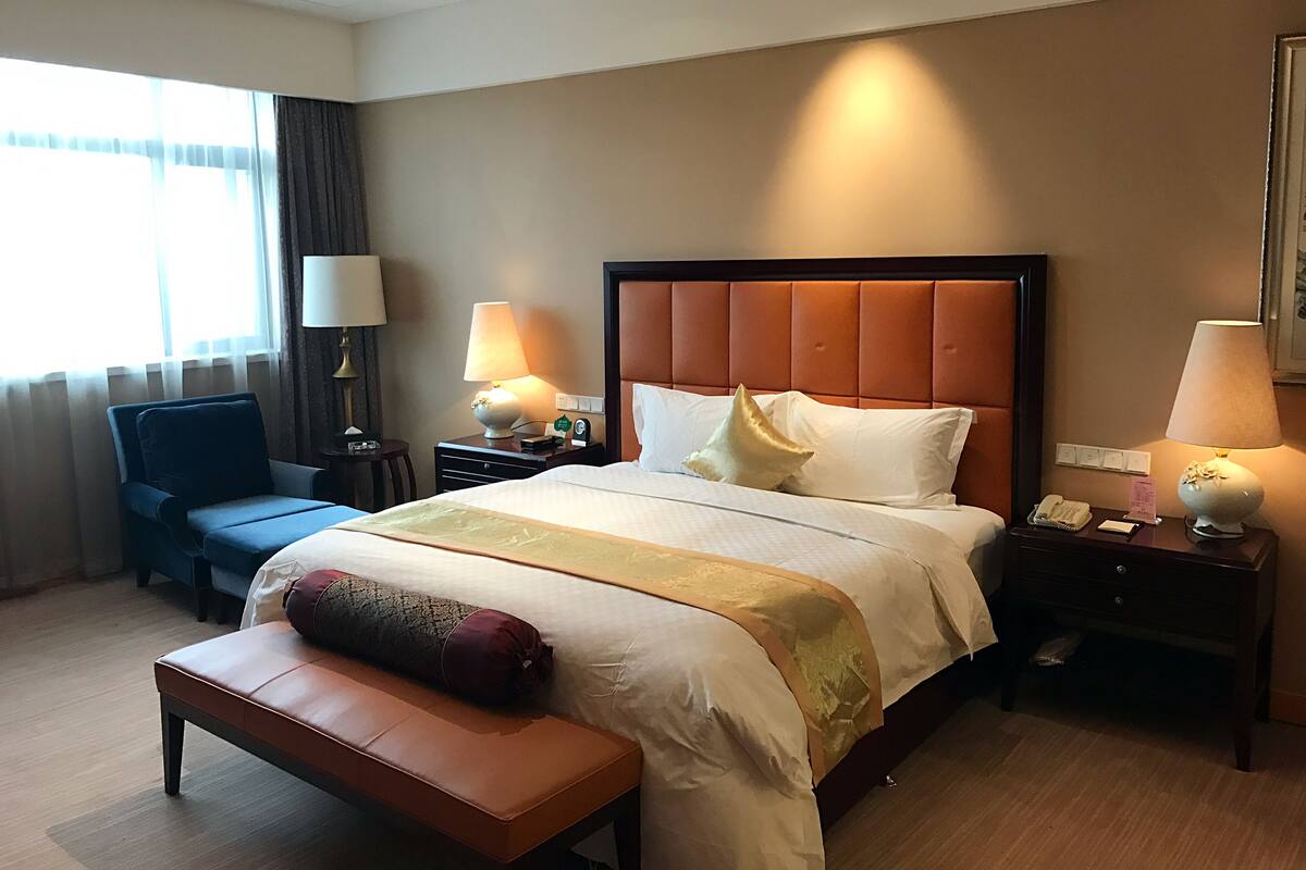 A hotel room with a large bed, a bedside table with a lamp, a chair, a bench, and a window with curtains.