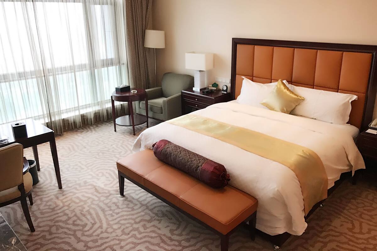A hotel room with a large bed, a bedside table, a chair, a desk, and a lamp.