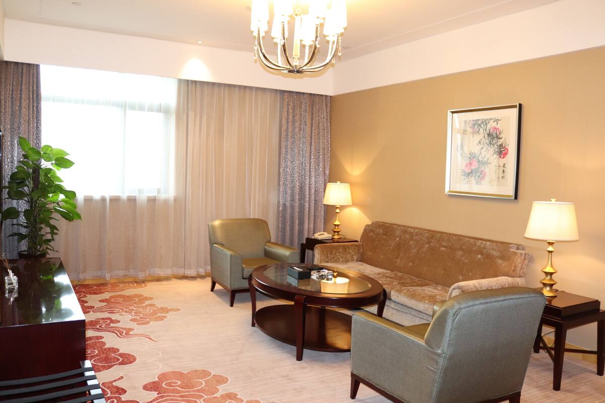 A hotel room with a sofa, two armchairs, a coffee table, a side table with a lamp, a potted plant, and a large window with curtains.