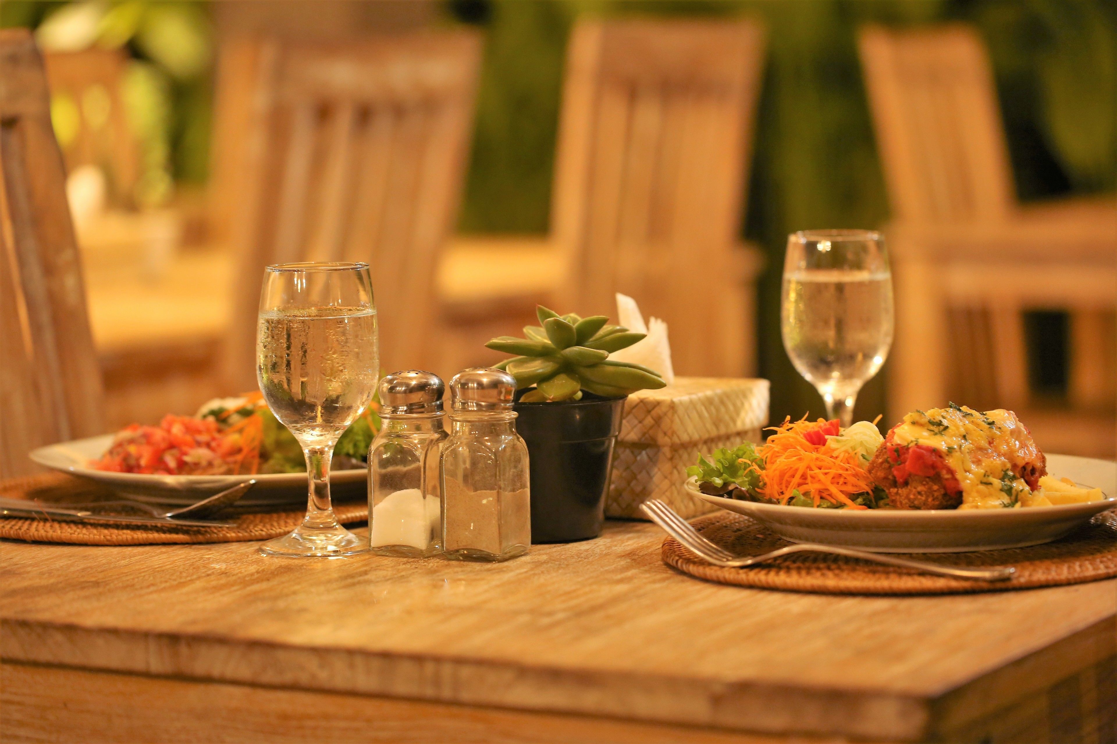 A wooden dining table with two plates of food, salt and pepper shakers, a small potted plant, and two glasses of water.