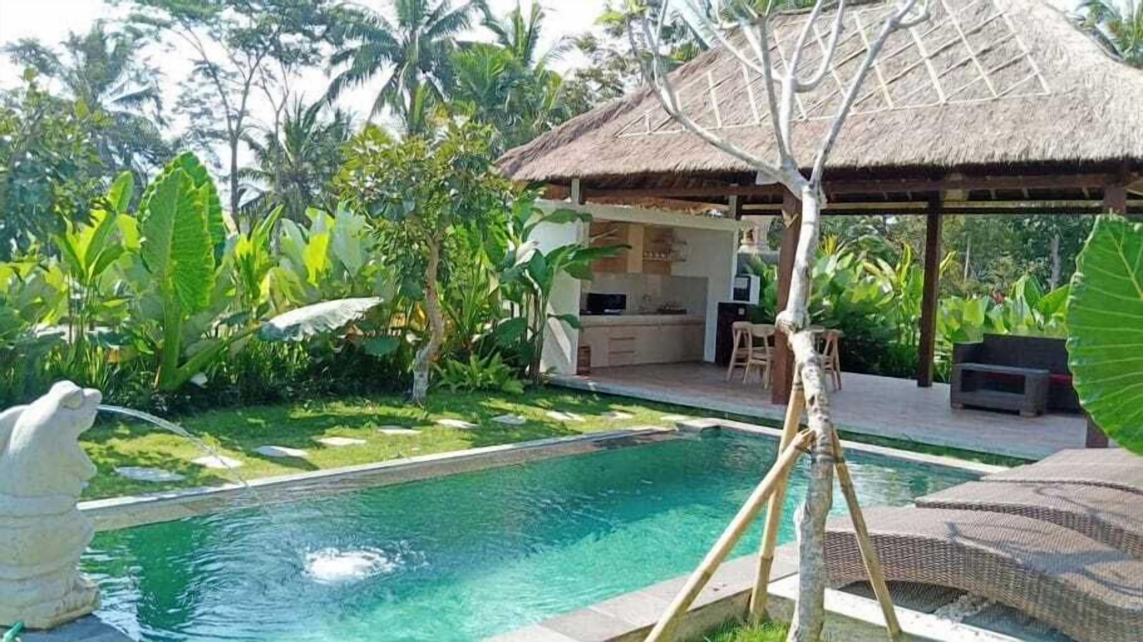 Una piscina con una estructura de techo de paja, rodeada de exuberante follaje y una estatua de piedra.