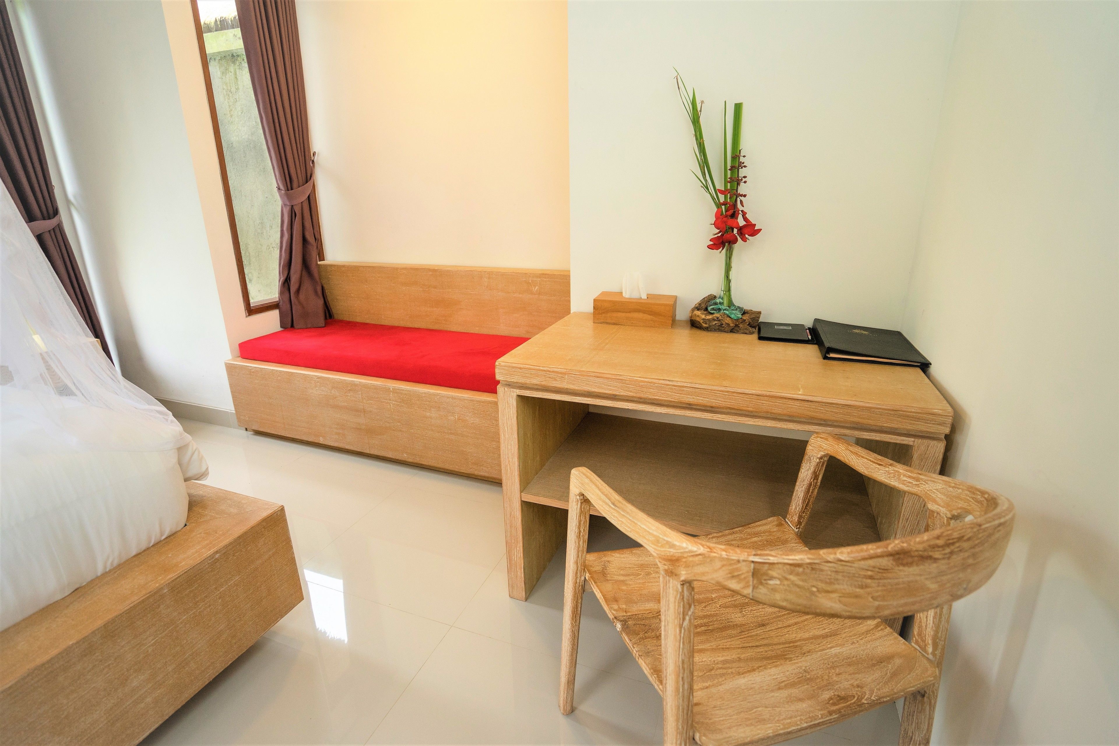 Habitación de hotel con una cama de madera, un sofá rojo, un escritorio de madera con una silla, un jarrón con flores y una mesita con un libro y una caja de pañuelos.