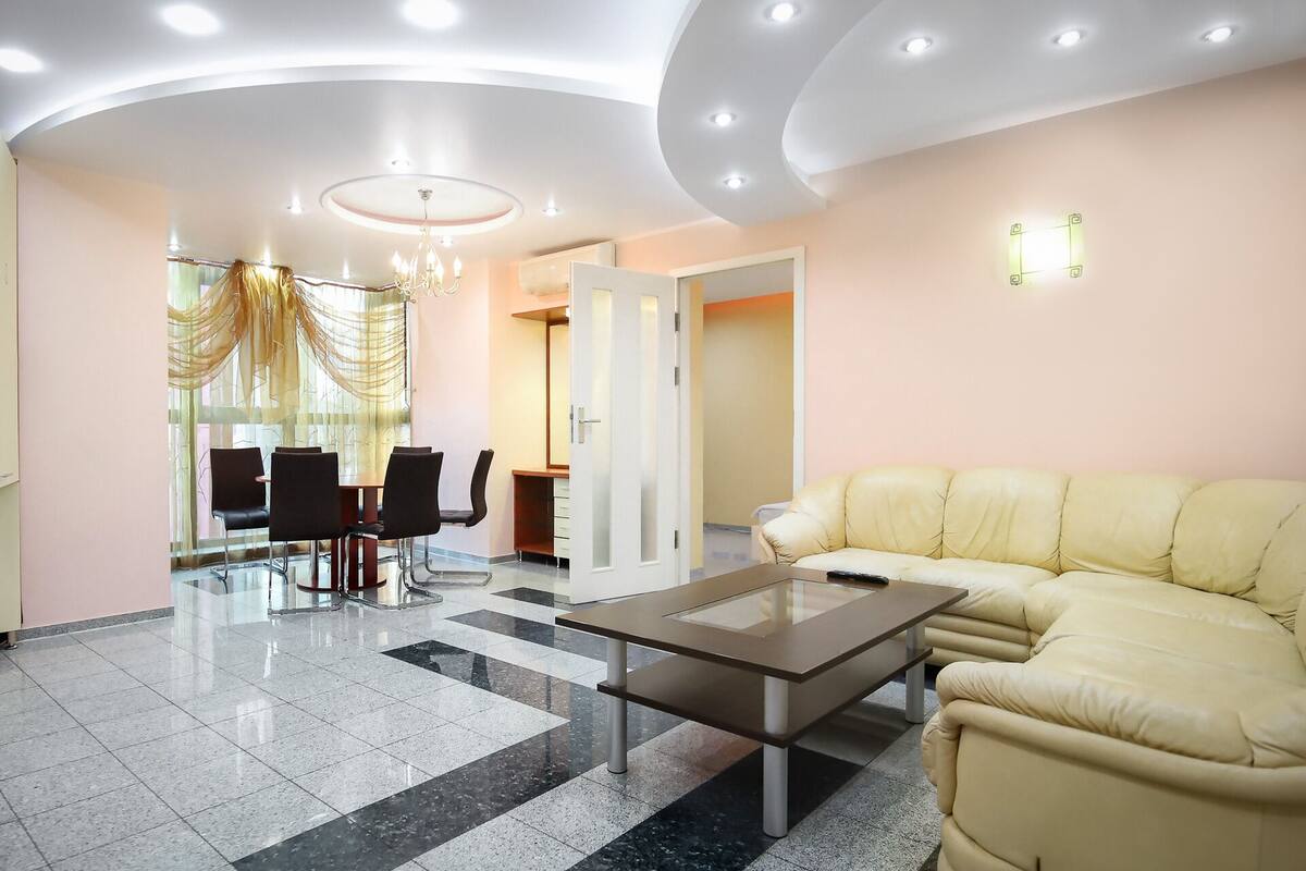 A modern living room with a beige sofa, a glass coffee table, and a dining area with chairs and a chandelier.