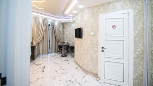A hallway with a marble floor, a white door with a no entry sign, a television mounted on the wall, and a small table with chairs. - Prestige Apart Hotel (Chisinau)