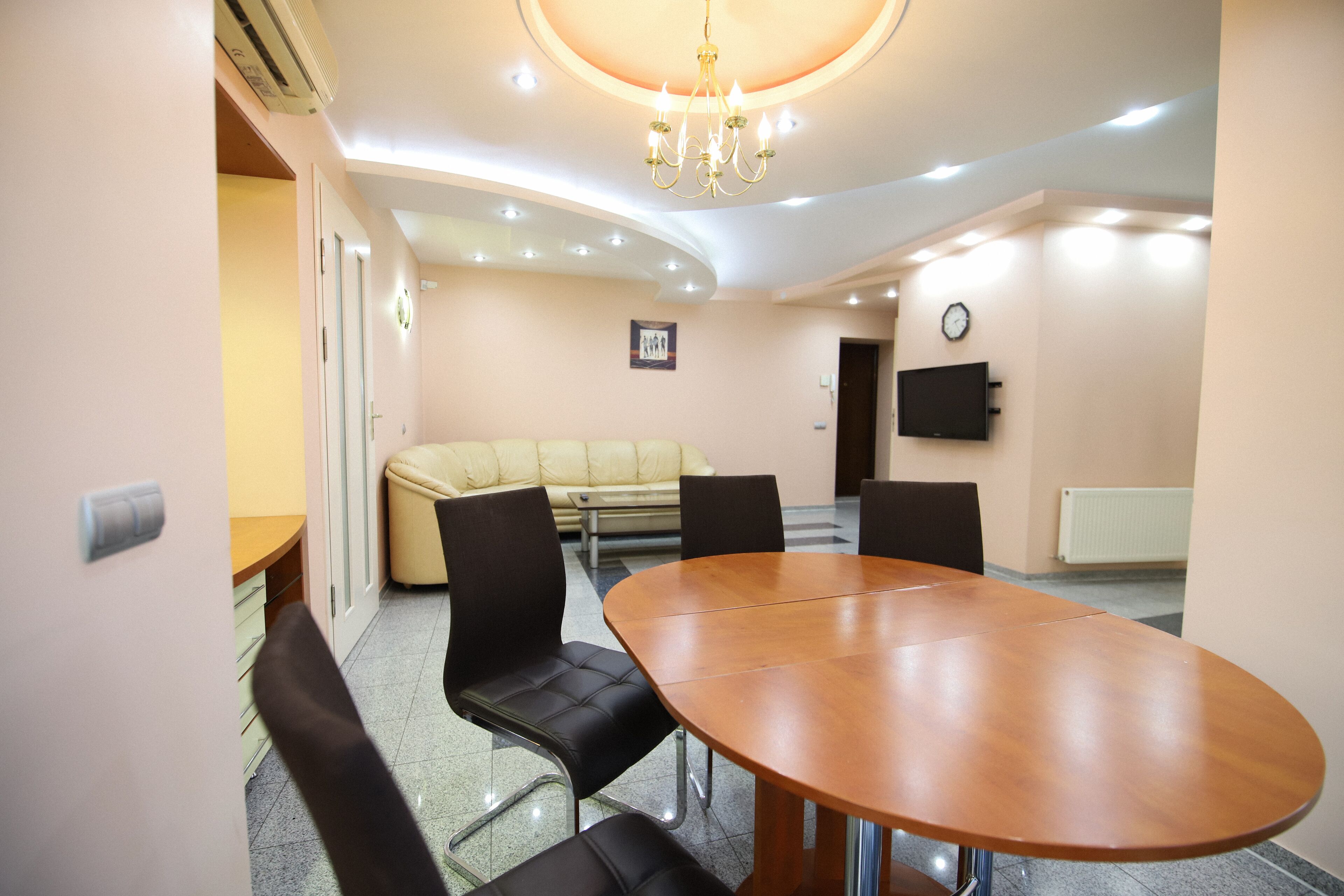 A conference room with a wooden table, black chairs, a beige sofa, a chandelier, recessed ceiling lights, a clock, and a TV on the wall.