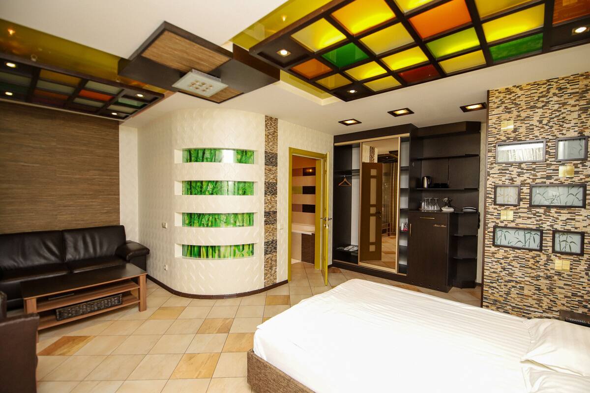A modern hotel room with a large bed, a sofa, a built-in wardrobe, and a bathroom with a glass shower.