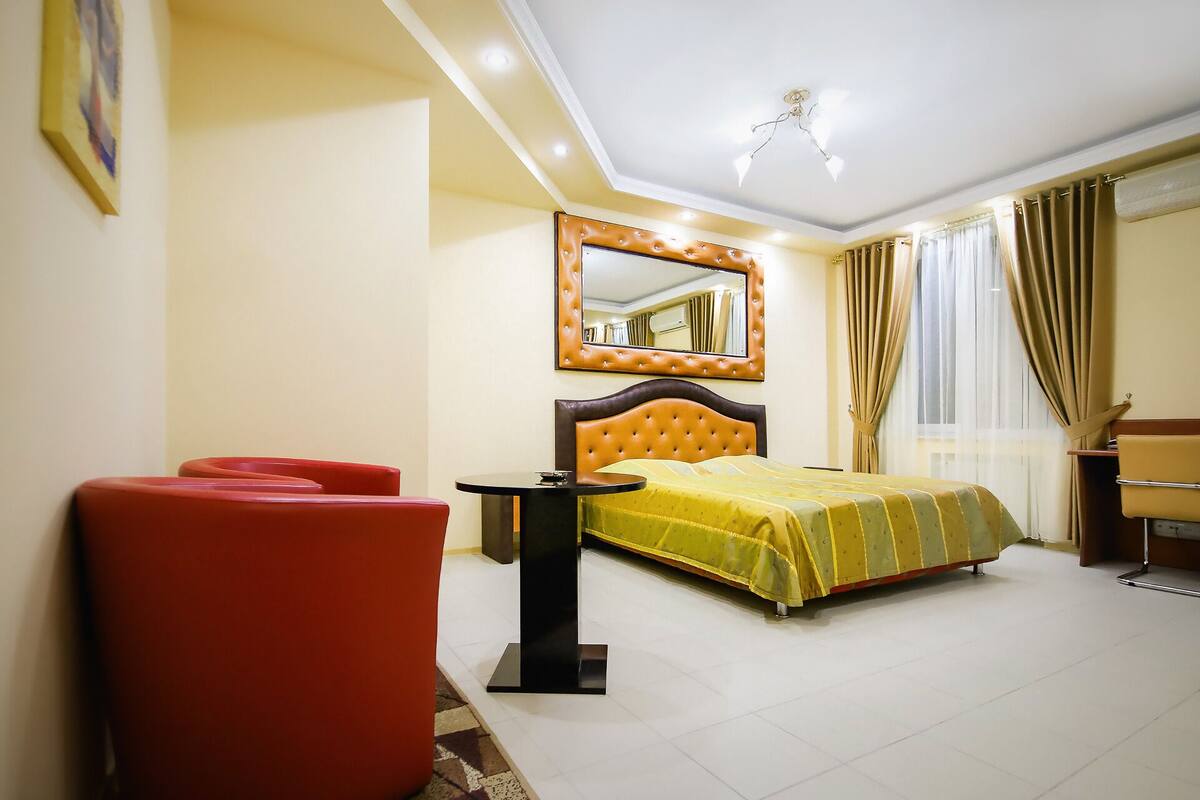 A hotel room with a bed, a red armchair, a small table, a mirror, and a window with curtains.