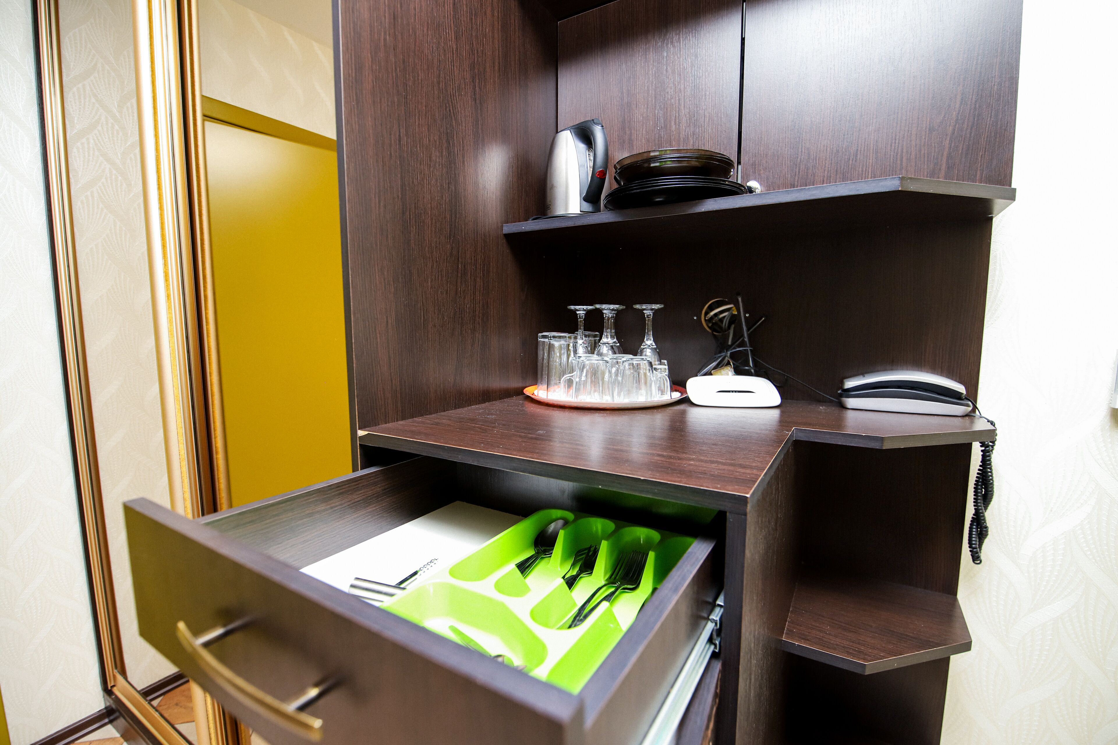 A hotel room with a wooden desk, a drawer with green utensils, a telephone, and a plate with glasses.