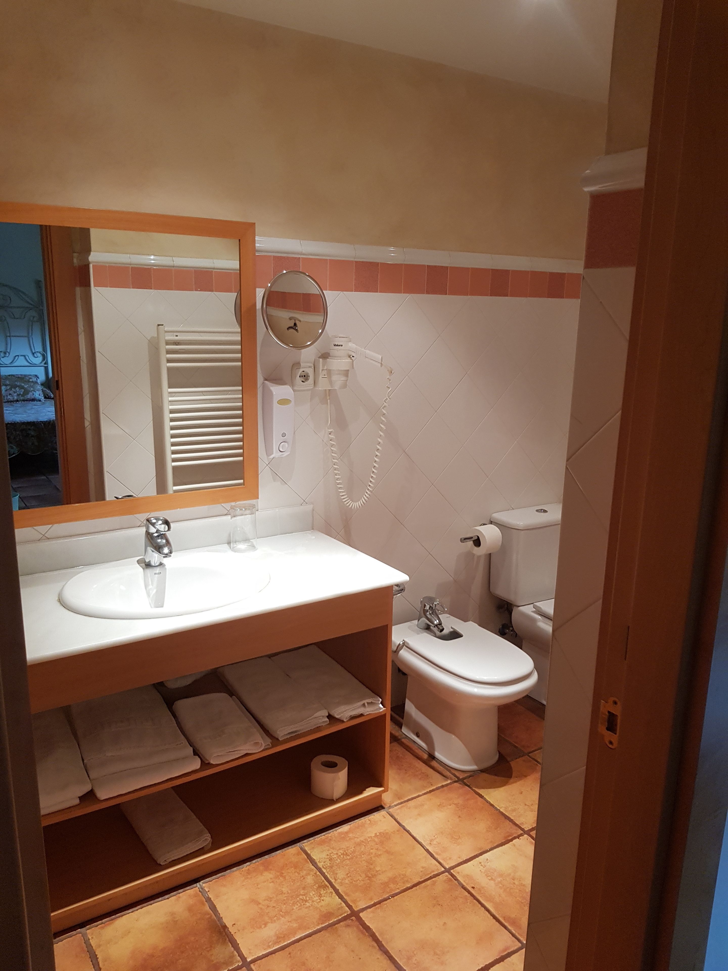 Ein Badezimmer mit einem weißen Waschbecken, Spiegel und Toilette. Ein Handtuchhalter mit gefalteten Handtüchern und ein Ablagebereich mit einer Toilettenpapierrolle.