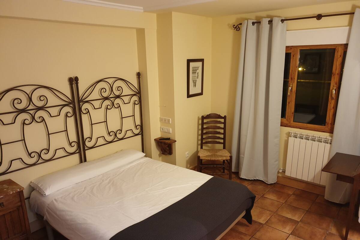 Un dormitorio con cama, una silla, ventana con cortinas, radiador y un armario de madera.