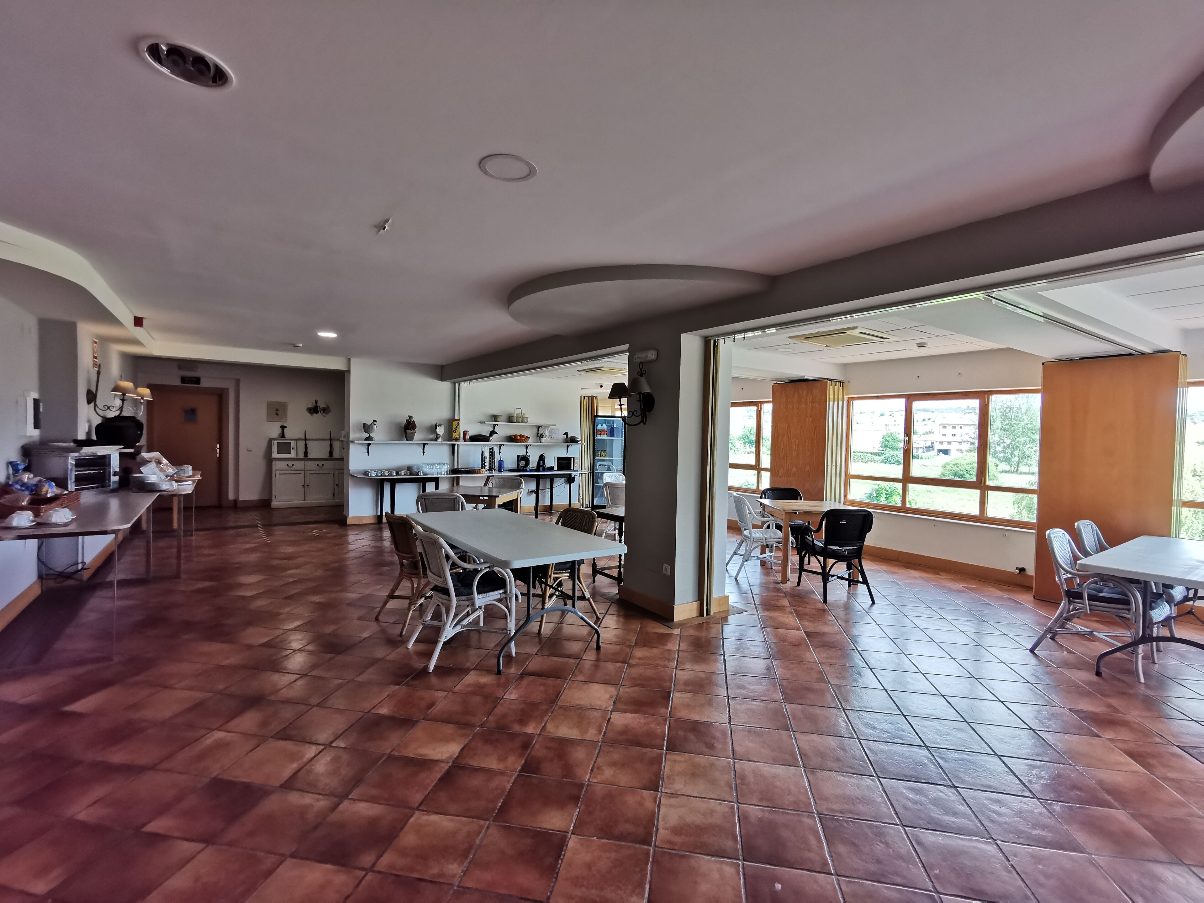 A spacious dining area with a long table and chairs, a kitchen area with shelves and appliances, and large windows providing natural light.