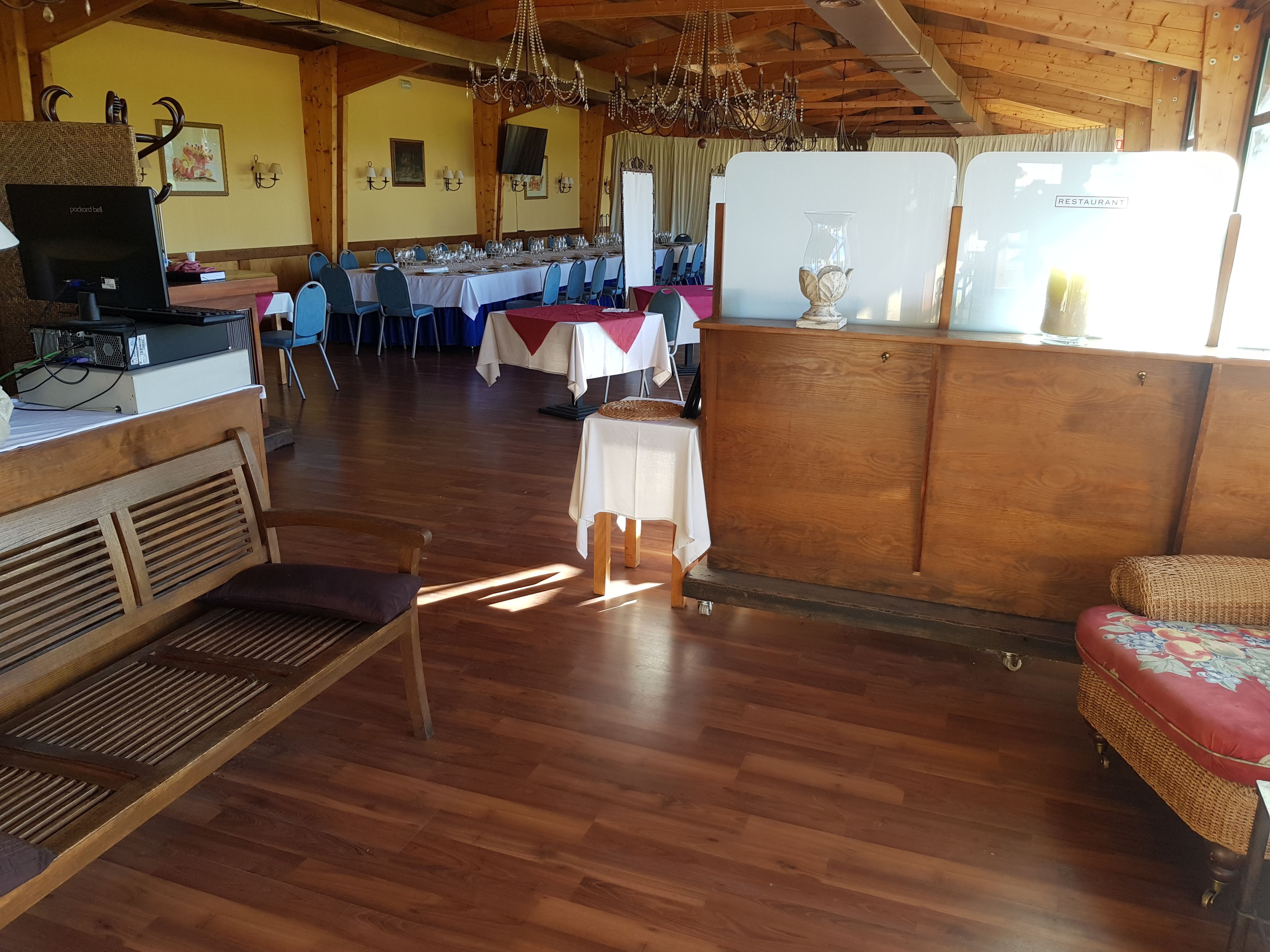 Un coin repas rustique avec des tables et des chaises en bois, un lustre et un comptoir en bois équipé d’un ordinateur et d’enceintes.