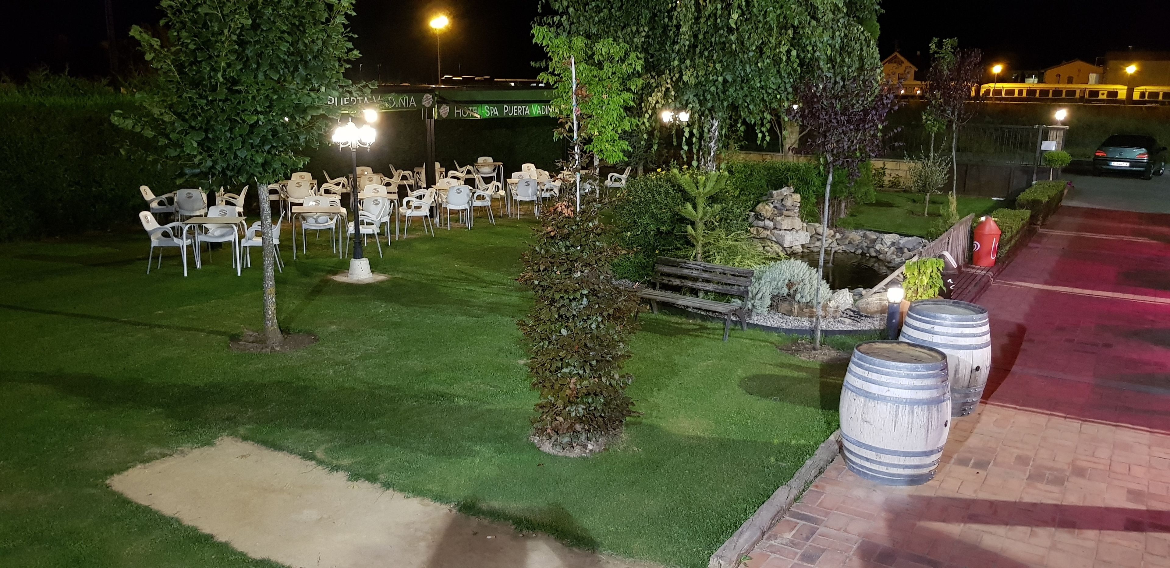 Un espacio al aire libre con mesas y sillas, rodeado de follaje y un muro de piedra.