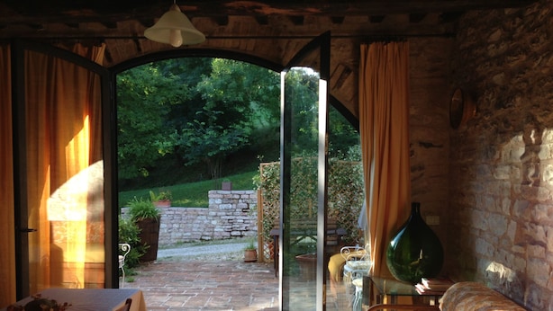Puerta abierta que da a un jardín con muro de piedra y follaje. Adentro, se ve una mesa con mantel blanco y una silla.