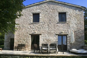 Un bâtiment en pierre doté de deux fenêtres, d’une porte et d’un patio comprenant une table et des chaises.