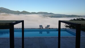 Una piscina con dos sillas de descanso frente a un paisaje montañoso parcialmente cubierto de niebla.