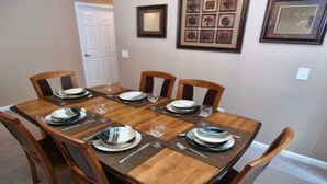 Mesa de jantar posta para seis lugares, com cadeiras de madeira, quadro na parede e uma porta ao fundo.