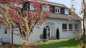 Une maison à deux étages, à l’extérieur blanc, avec un toit recouvert de tuiles et un petit jardin devant, agrémenté d’un arbre et d’un banc.