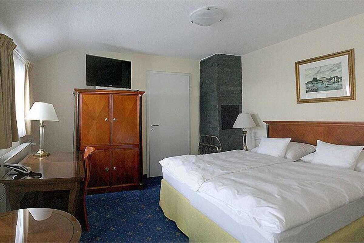 A hotel room with a bed, a desk with a lamp, a wardrobe, a television, and a framed picture on the wall.