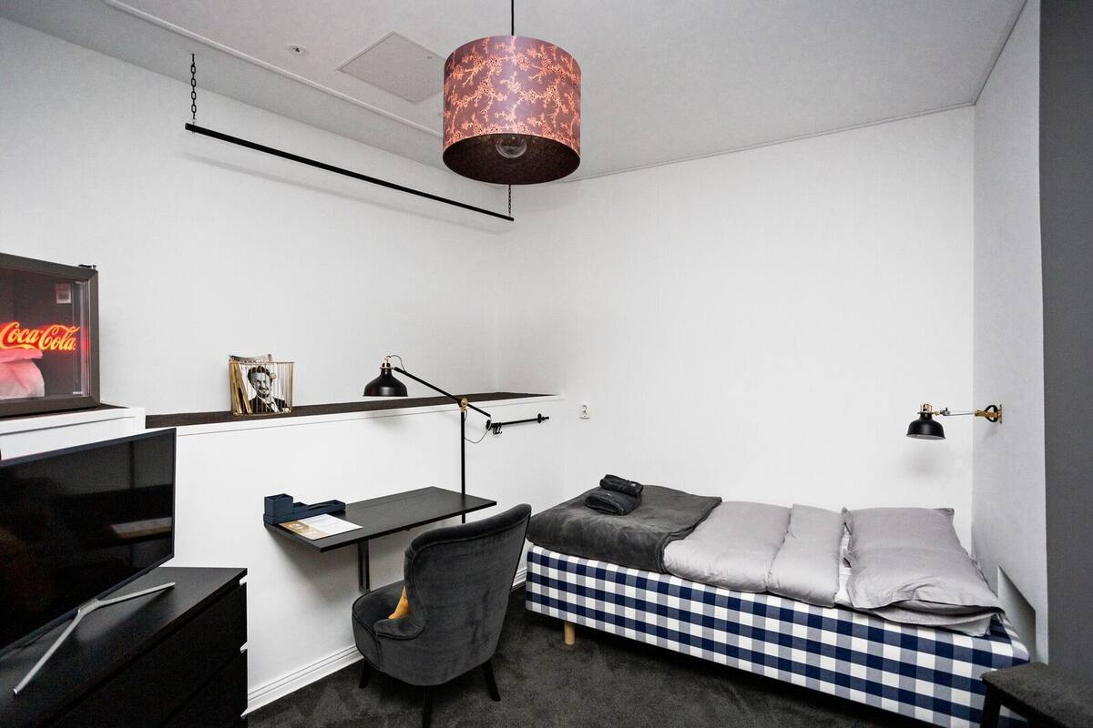 Una habitación pequeña con cama, escritorio, silla y una lámpara colgante.