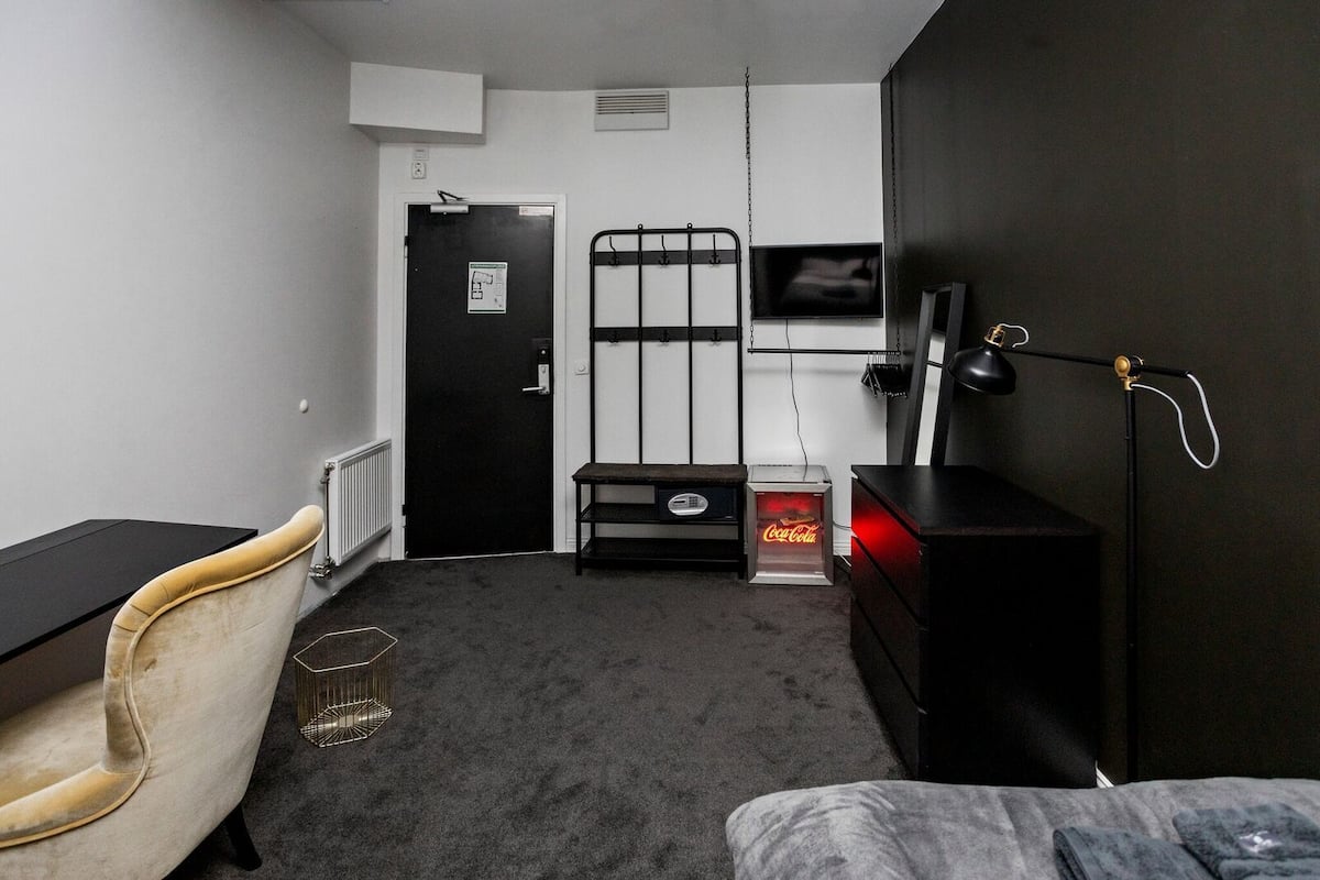 Una habitación pequeña y poco iluminada con una cama, un escritorio, una silla, un radiador, una puerta, un televisor y una máquina expendedora de Coca-Cola.