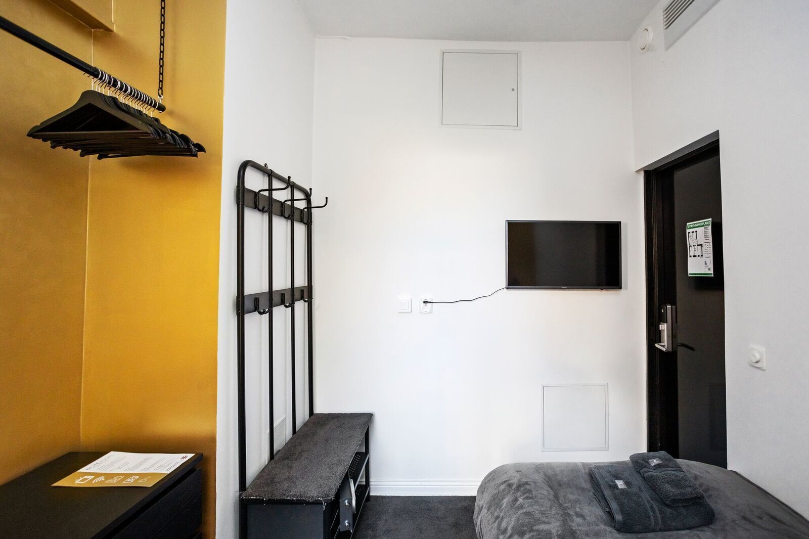 Una habitación pequeña con cama, un banco, televisor montado en la pared y perchero.