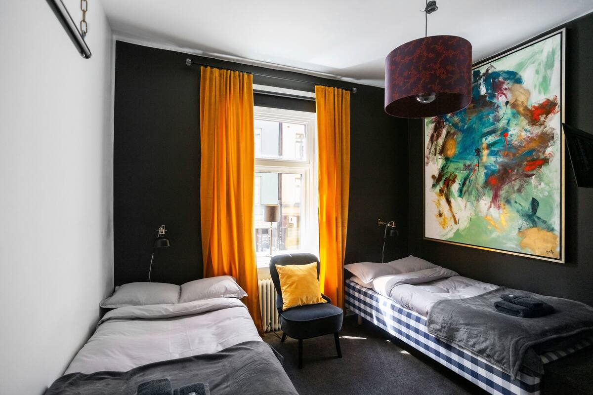 Una habitación de hotel con dos camas, una silla, un cuadro abstracto grande y una ventana con cortinas.