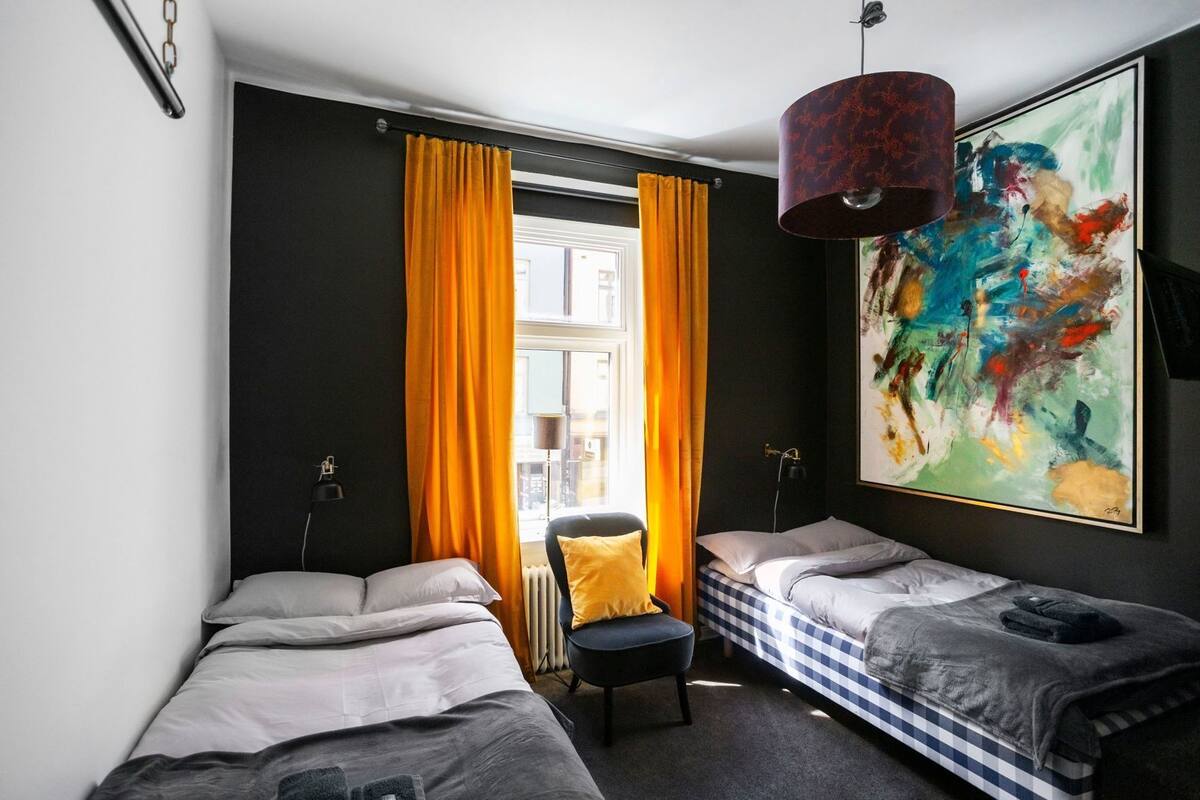 Un dormitorio con dos camas, una silla, un cuadro abstracto grande y un ventanal con cortinas.