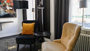 A modern living room with a beige armchair, a black sofa, a small black table, and a floor lamp. There is a large abstract painting on the wall and a window with curtains. - Eight Rooms (Stockholm)