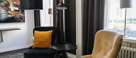 A modern living room with a beige armchair, a black sofa, a small black table, and a floor lamp. There is a large abstract painting on the wall and a window with curtains.