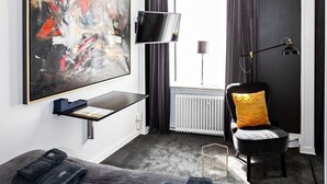 A modern hotel room with a large abstract painting, a flat-screen TV, a bed with dark linens, a chair with a yellow pillow, and a large window with curtains. - Eight Rooms (Stockholm)