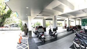 A covered parking area with multiple motorcycles parked, a large tree, and a white van in the background. - Tree House Apartment Songkhla (Songkhla)