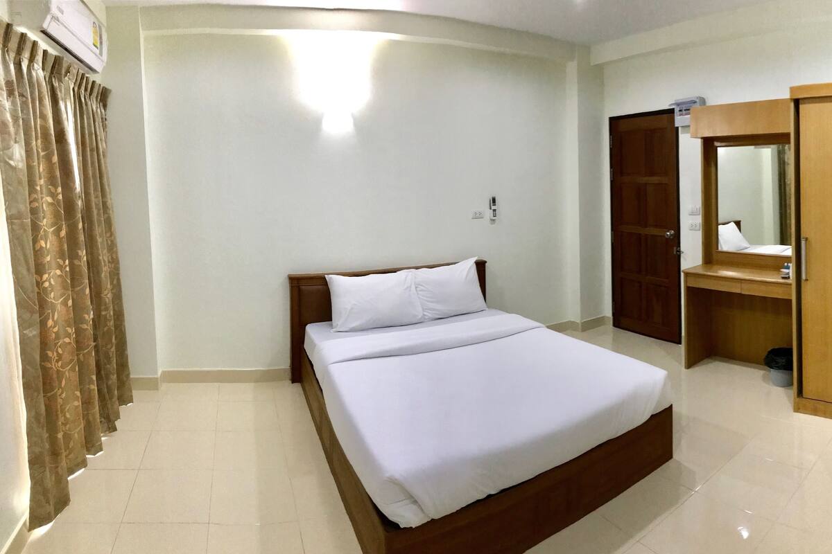A hotel room with a bed, a wooden wardrobe, a small bathroom with a mirror, and a ceiling-mounted air conditioning unit.