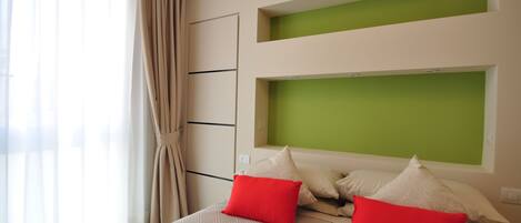 Una habitación de hotel moderna con una cama grande, ropa de cama color beige y almohadas rojas. Hay paredes de acento verde y una ventana con cortinas translúcidas.