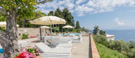 Un espace aménagé au bord de la piscine, avec des chaises longues et des parasols, une piscine et une vue sur la mer.