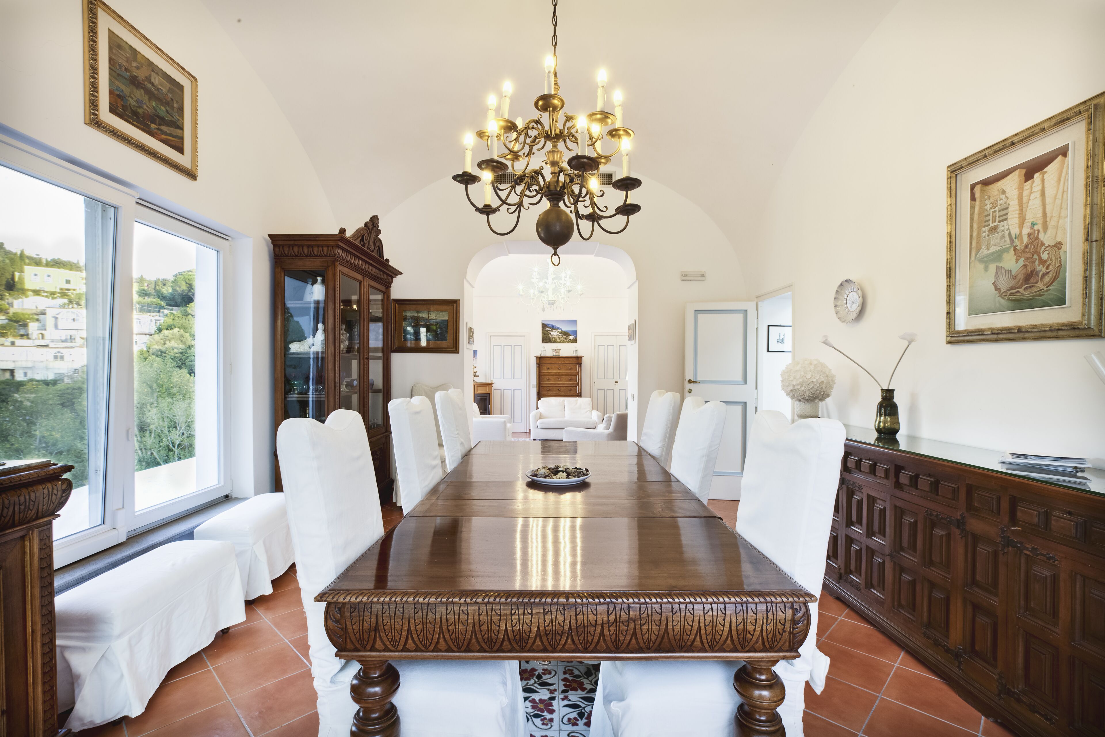 A dining room with a large wooden table, white upholstered chairs, a chandelier, and a view through large windows.